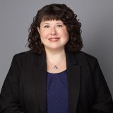 Woman with dark curly hair in black blazer and navy top smiling.