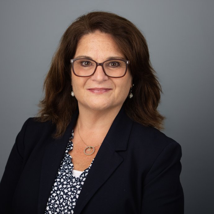 Headshot of a woman wearing a dark blazer and patterned blouse against a gray background.